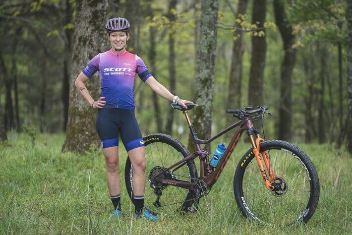 Kolesarka pozira ob kolesu, na travnati jasi v gozdu.