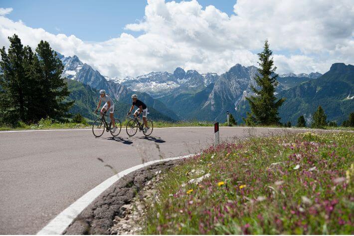 Ženska in moški kolesarita po asfaltirani cesti v klanec, v ozadju so gore.